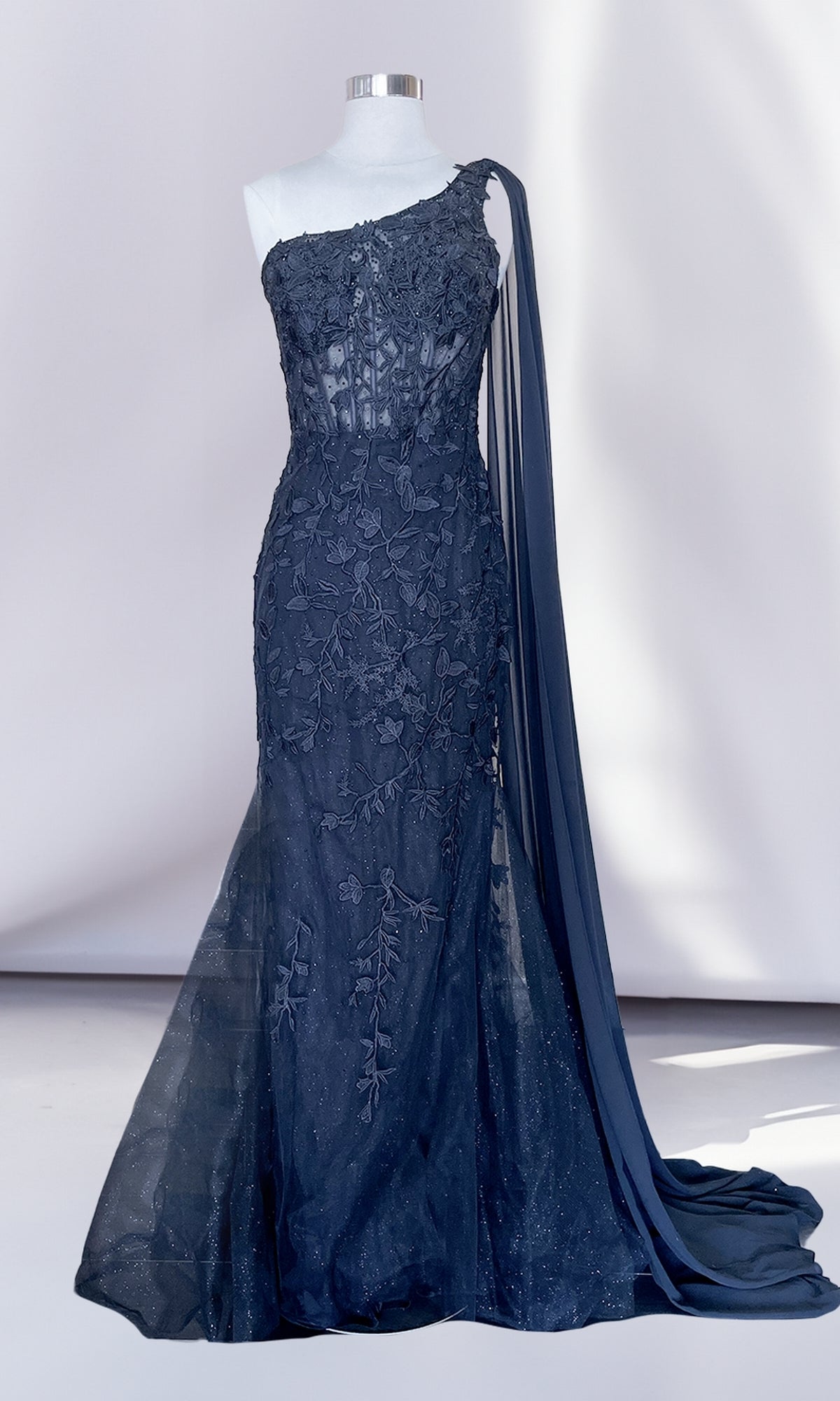 The One-Shoulder Long Lace Mermaid Prom Dress 7048 in navy blue features floral embroidery, sheer detailing, and a long draped sash, elegantly showcased on a dress form.
